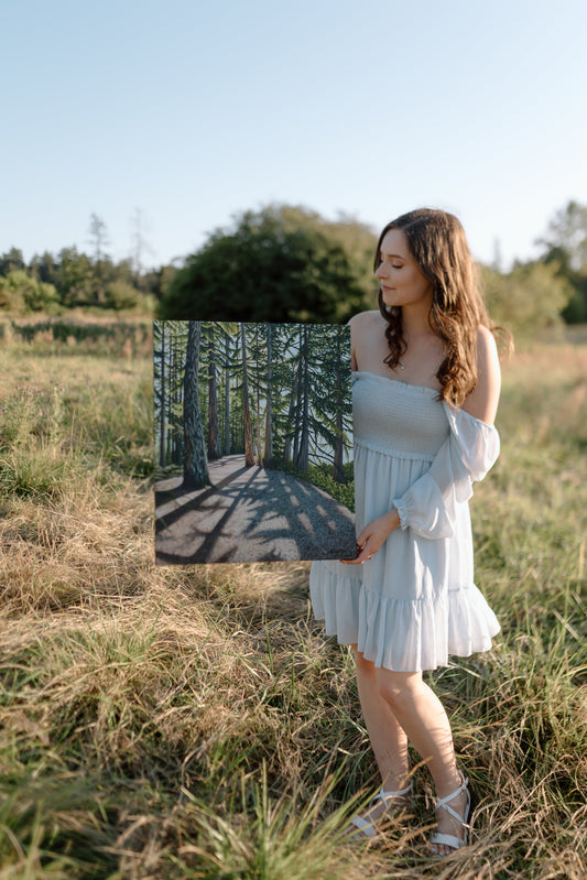 The artist, Meaghan Crow holds her painting "Elk Lake Sunlit Trail" in a grassy field. The painting shows sunlight streaming through the trees creating shadows on the trail below.